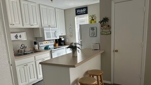 A kitchen with white cabinets, a central island with stools, a coffee maker, and a refrigerator. - 2 Br 1 Bath Condo in Gulfshores, AL, 2 min walk to the Beach, Pool and Fishing (Gulf Shores)