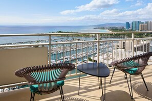 Un balcon avec deux fauteuils en osier et une petite table, d’où l’on peut admirer une marina avec de nombreux bateaux et, au loin, une plage.