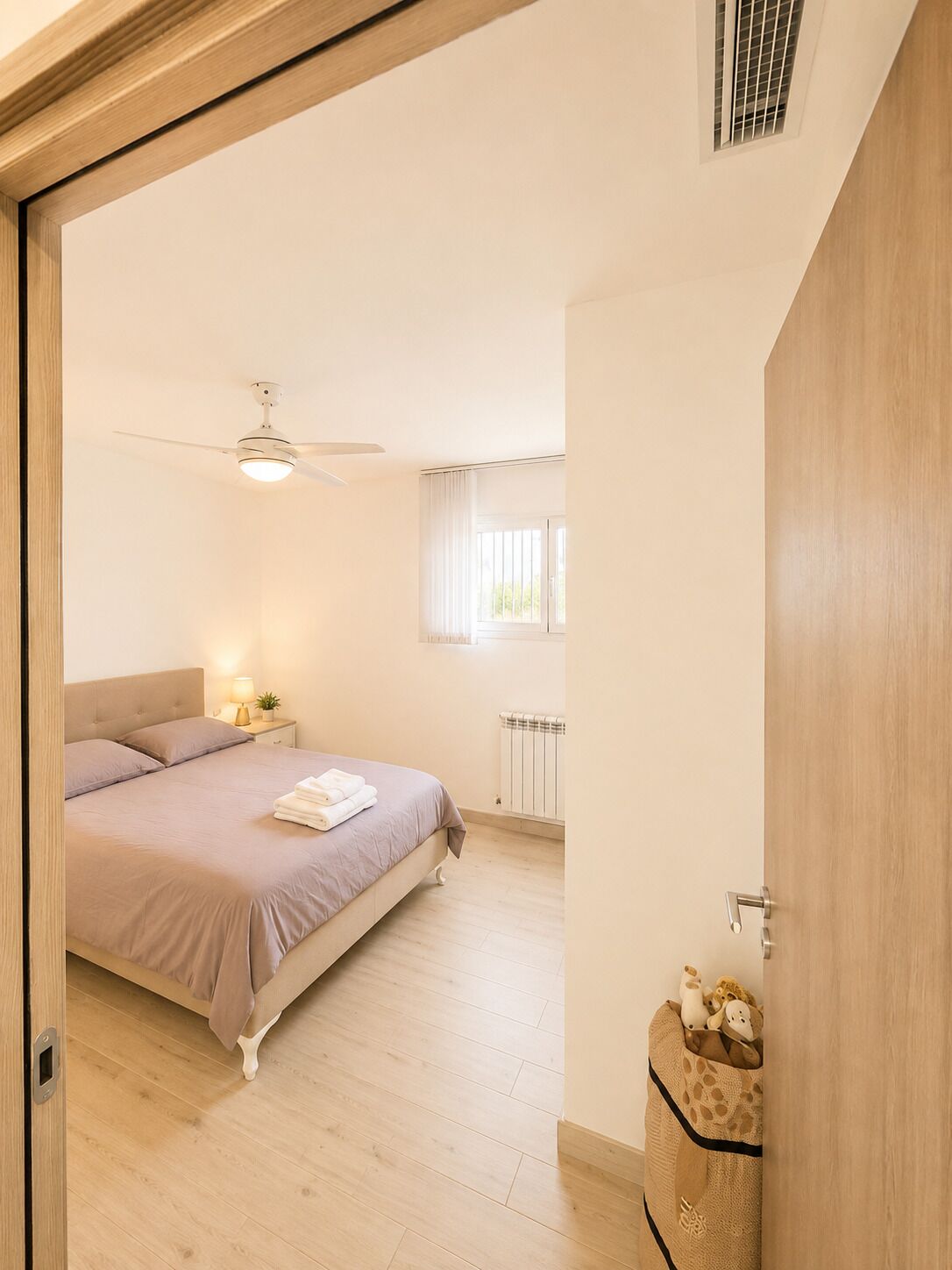 Un dormitori amb un llit, una tauleta de nit, un ventilador de sostre, una finestra amb cortines i un radiador.