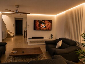 A modern living room with a flat-screen TV mounted on the wall, a ceiling fan, a staircase leading to another level, and a coffee table with remote controls.