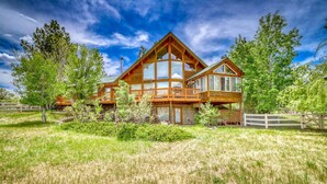 A large wooden cabin with multiple decks and large windows, surrounded by a grassy field and trees.