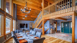A spacious dining area with a high ceiling and wooden beams, a staircase leading to an upper level, and a cozy seating area with a fireplace.