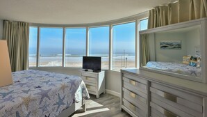 A hotel room with a large bay window, a bed with a star-patterned bedspread, a bedside table with a television, and a dresser with a mirror. - LUXURY OCEANFRONT 2BR Condo, Pool & Hot Tub, Game Rooms, Great Views (Daytona Beach Shores)