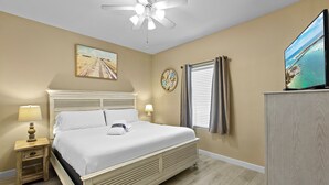 Une chambre à coucher avec un lit, des lampes de chevet, un ventilateur de plafond, un tableau, une fenêtre avec des rideaux et un téléviseur posé sur un meuble.