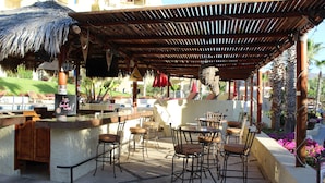 Zona de bar al aire libre con techo de paja, taburetes y una piscina al fondo.