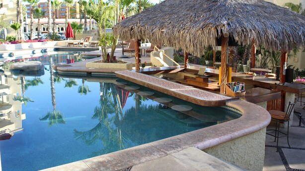 Un área de piscina con una estructura de techo de paja, sillas de descanso y palmeras.