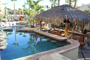 A pool area with a thatched-roof structure, lounge chairs, and palm trees. - Misiones del Cabo 4206. 1BR Completely Remodeled Ocean View Condo! (Cabo San Lucas)