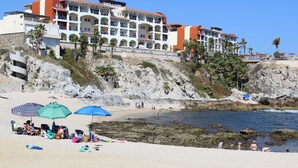 Un hotel frente a la playa con varios balcones, una playa de arena y acantilados rocosos.
