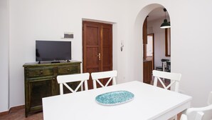 Een eetgedeelte met een witte tafel, vier witte stoelen, een blauw bord met patroon, een televisie op een houten kast en een houten deur.