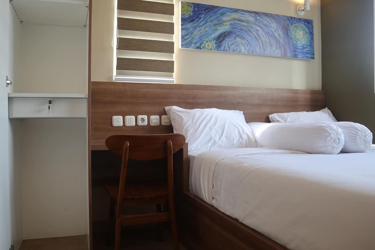 A bedroom with a wooden headboard, a bed with white linens, a chair, a painting on the wall, and a window with blinds.