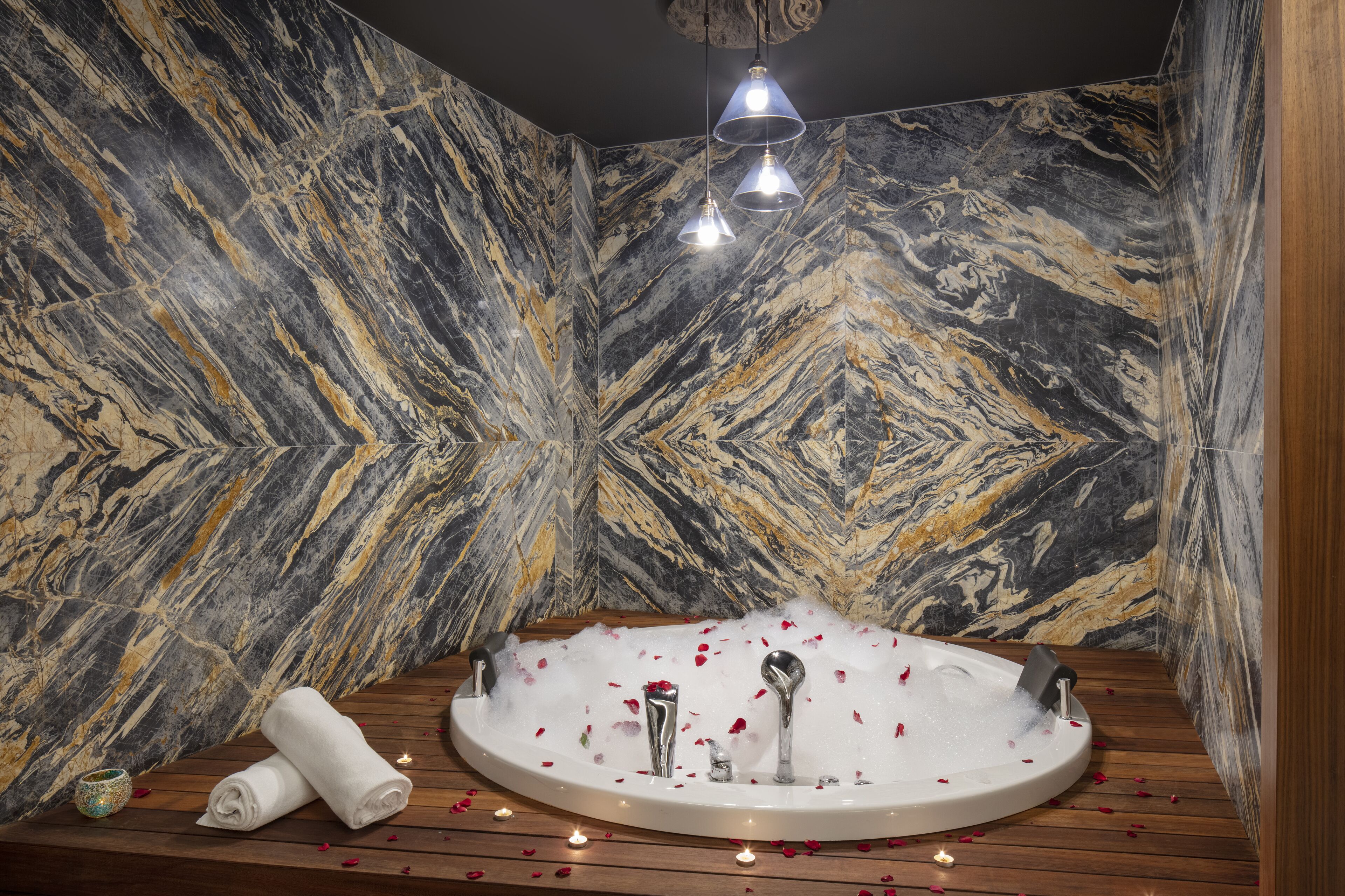 A spa room with a marble wall, a wooden floor, a round bathtub with rose petals, and three pendant lights.