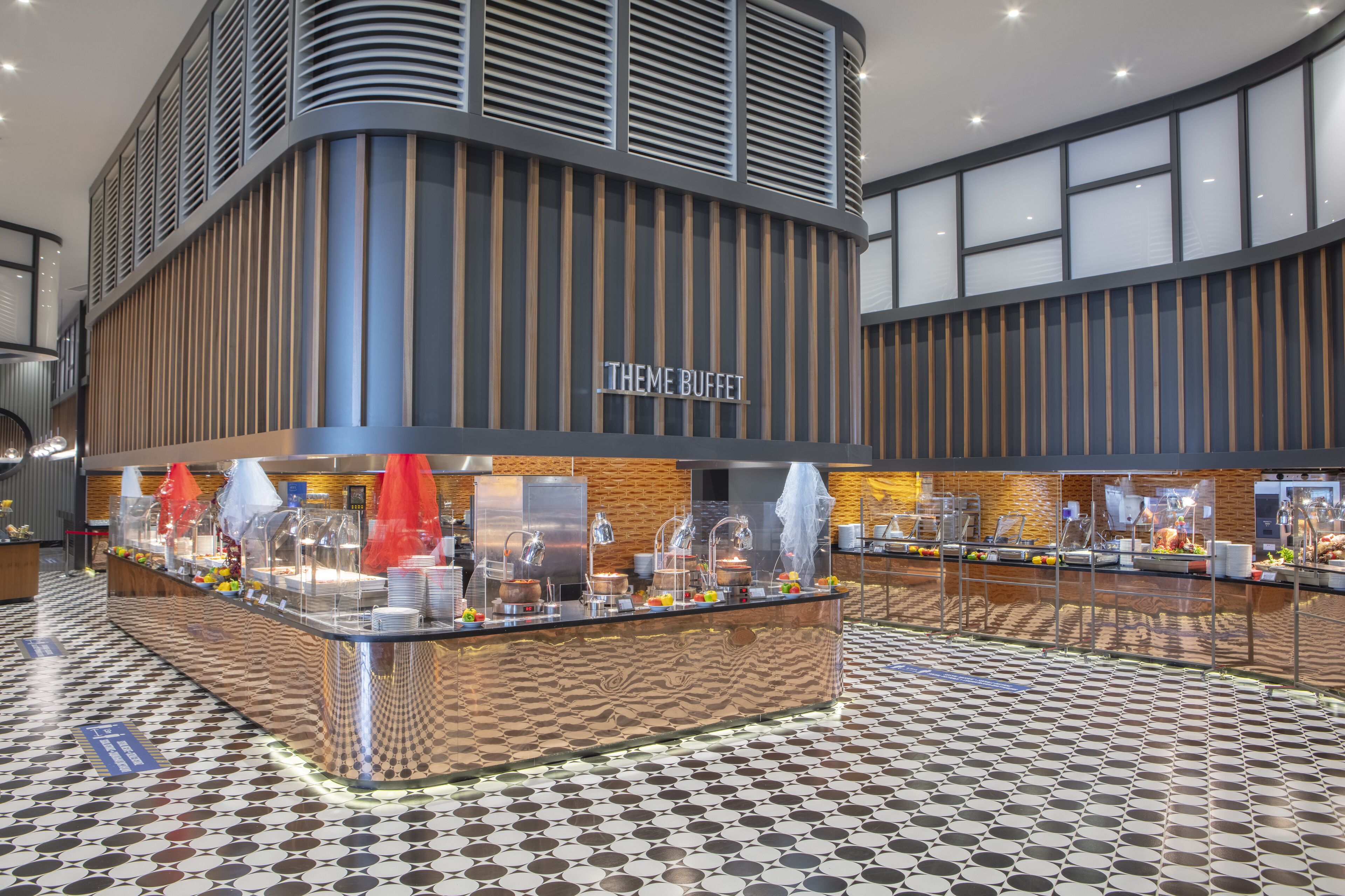 A buffet area with a modern design, featuring a central counter with food displays and a sign that reads "THEME BUFFET".