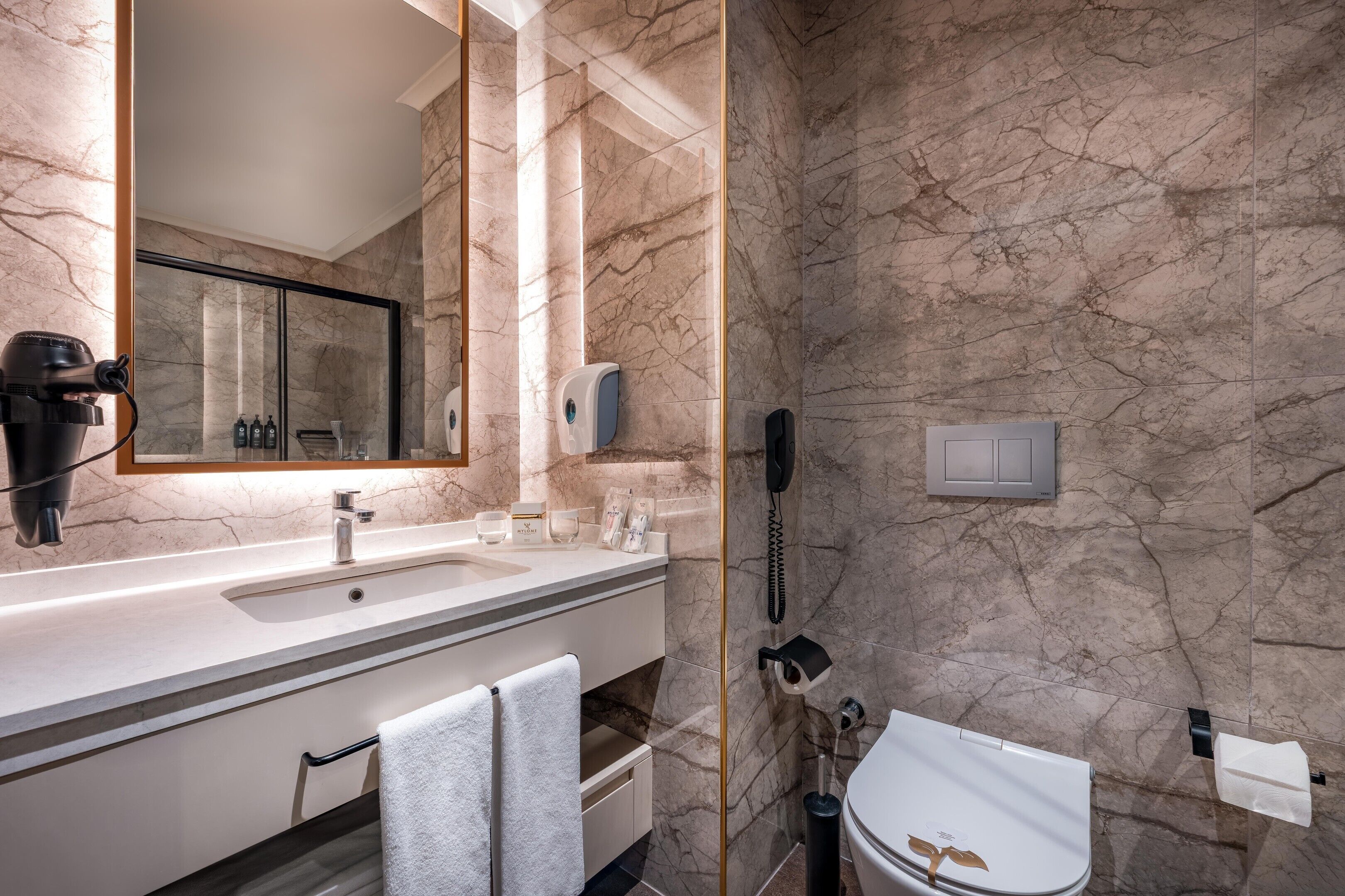 A bathroom with a marble wall, a sink with a mirror, a hairdryer, a phone, and a toilet.