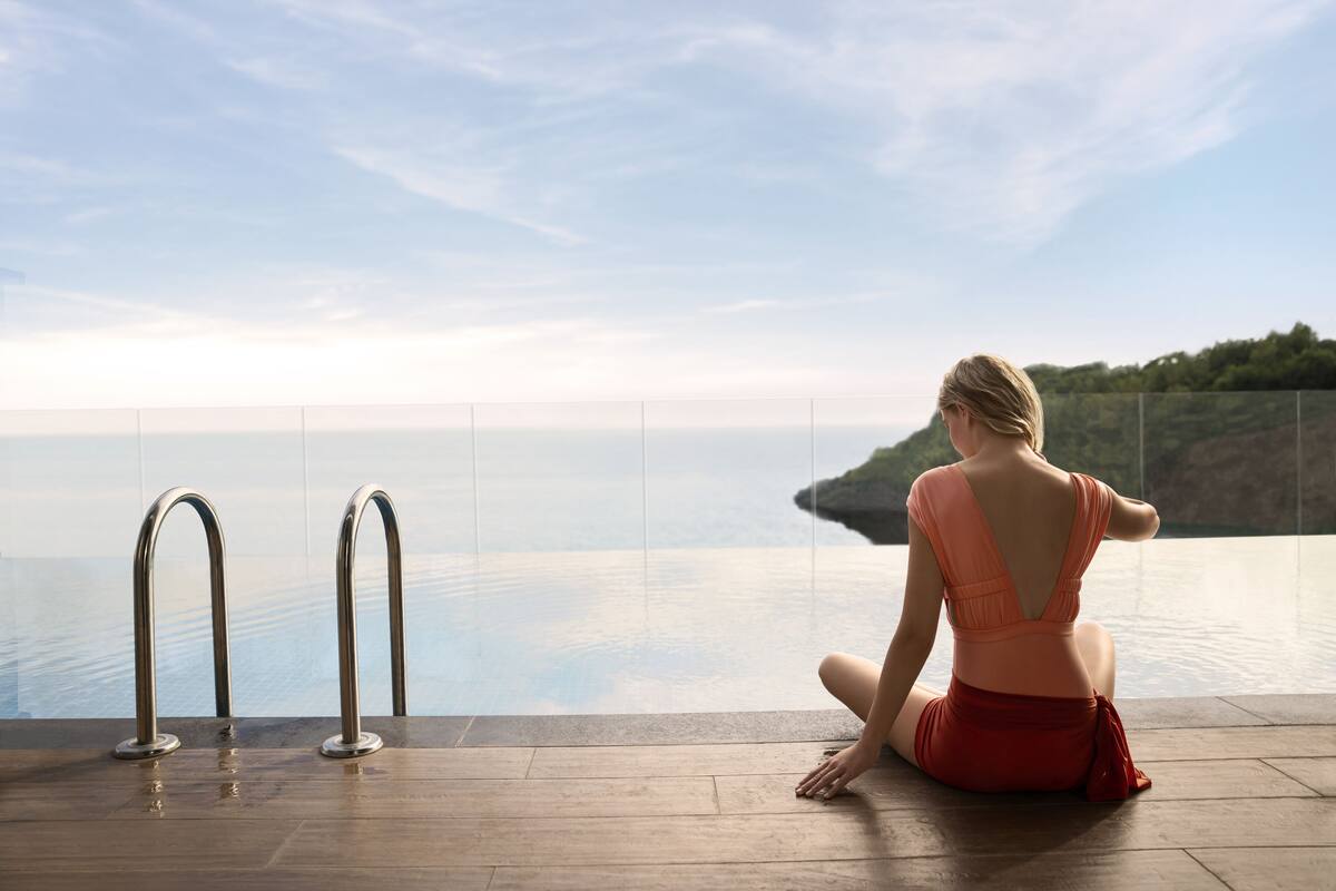 Eine Frau sitzt am Pool mit Blick auf das Meer und eine Klippe.