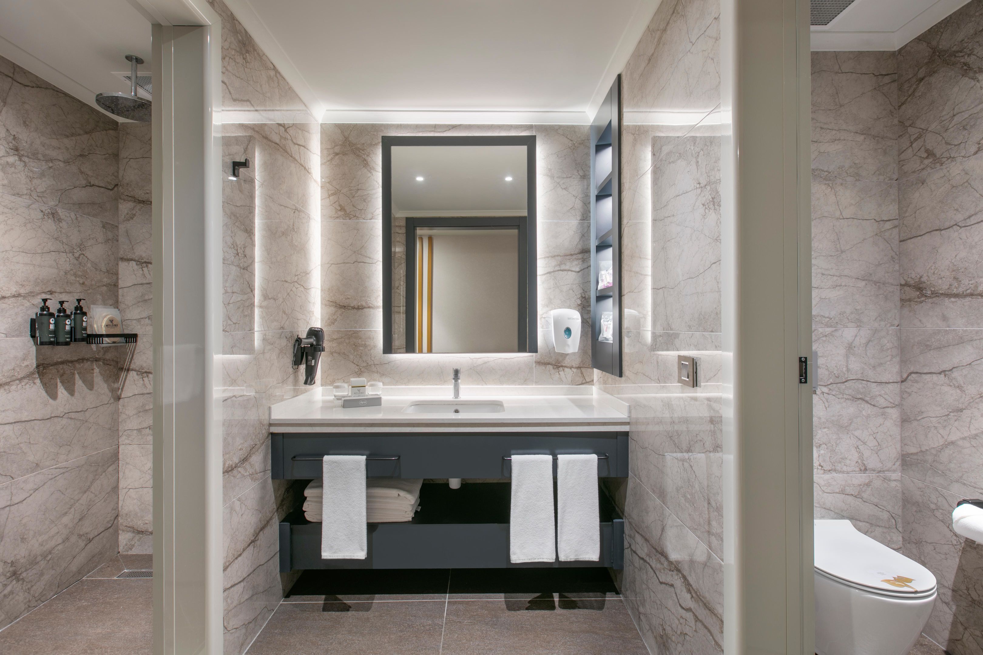 A modern bathroom with a marble wall, a large mirror, a sink, a toilet, and a shower area.