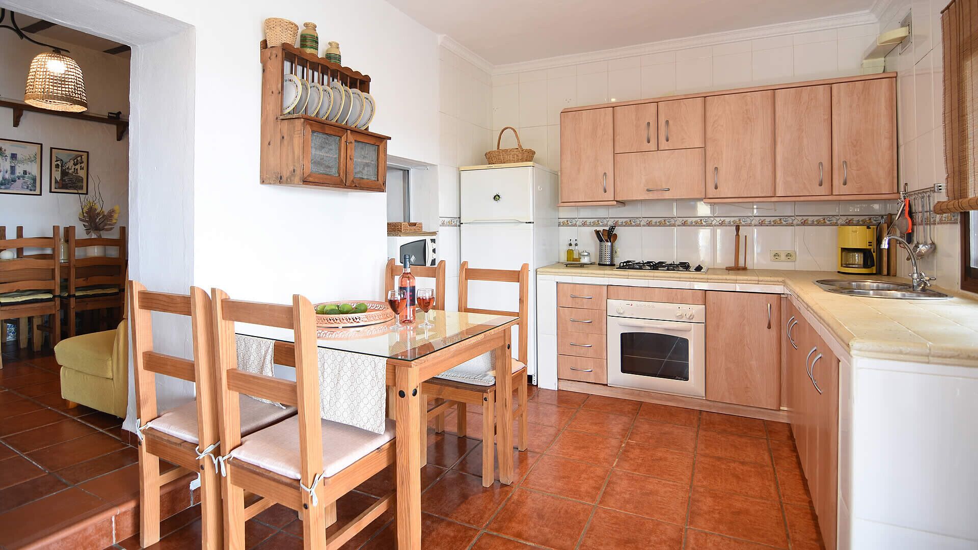 Een kleine keuken met houten kastjes, een eettafel met stoelen, een koelkast en een koffieapparaat.