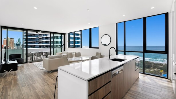 A modern kitchen with a large island, a dining area with a table and chairs, and a view of the ocean through large windows. - Koko Broadbeach (Broadbeach)