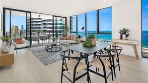A modern living room with a large window overlooking the sea, a dining area with round tables, and a cozy seating area with a sofa and armchairs. - Koko Broadbeach (Broadbeach)