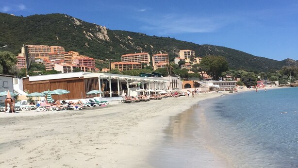 Una playa con aguas cristalinas, una orilla arenosa y edificios al fondo.