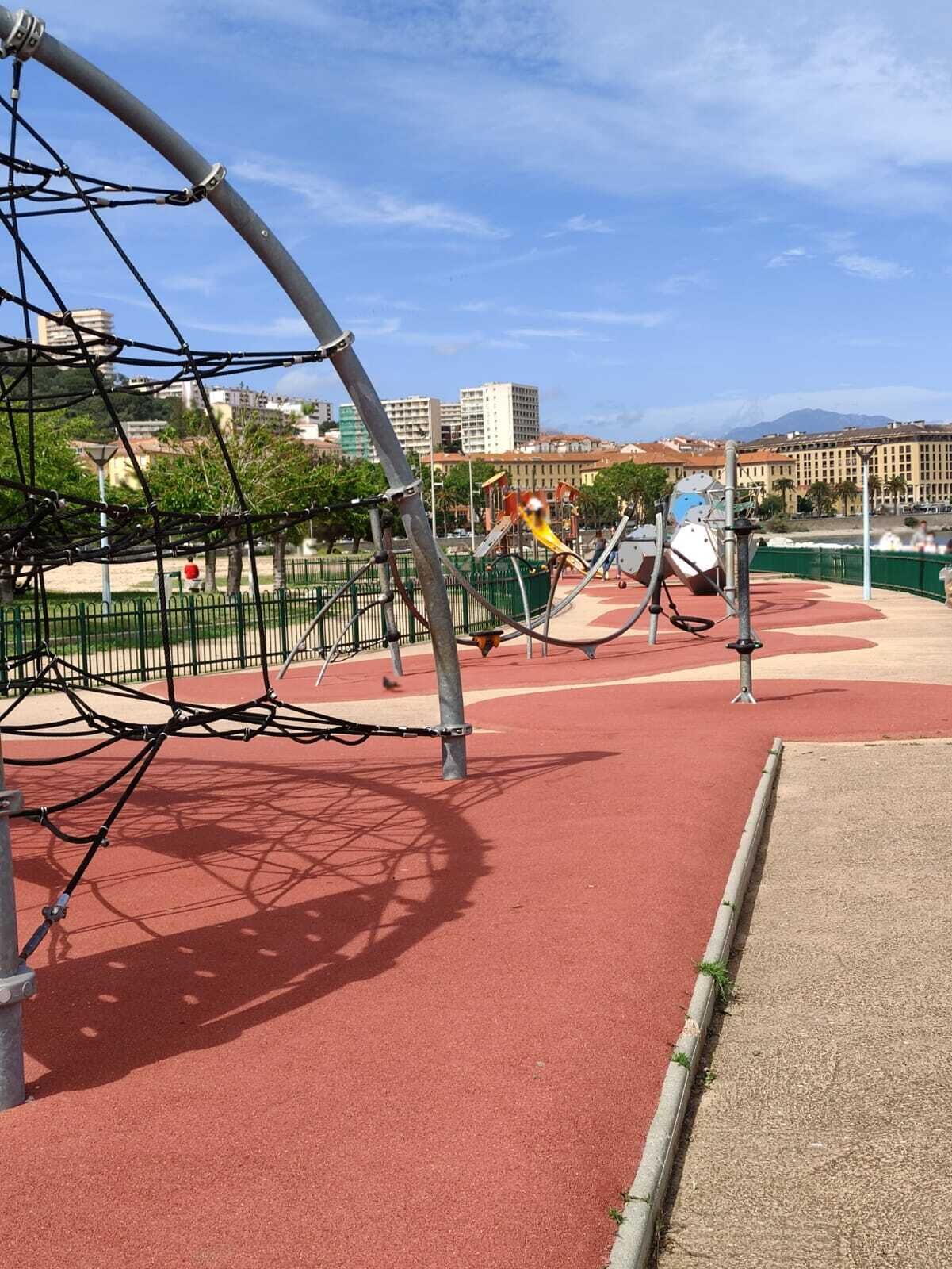 Zona de jocs amb estructures per escalar i tobogans, envoltada d'una pista de running vermella i una tanca verda.