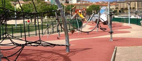 Zona de jocs amb estructures per escalar i tobogans, envoltada d'una pista de running vermella i una tanca verda.