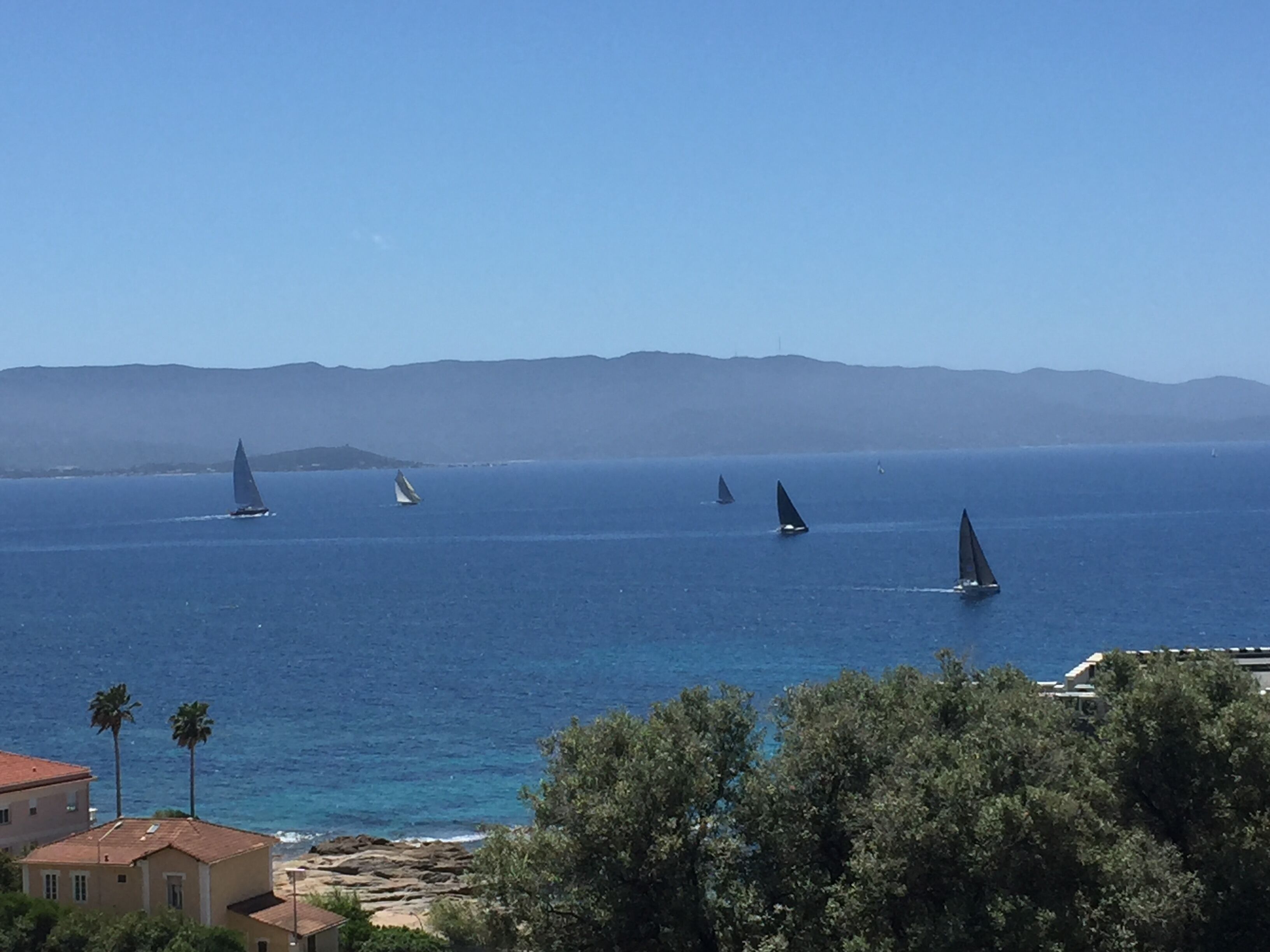 A coastal view with sailboats on the sea, a rocky shoreline, and buildings in the foreground.
