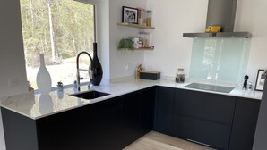 A modern kitchen with black cabinets, a white countertop, and a window providing a view of trees. - Villa (Talloires-Montmin)