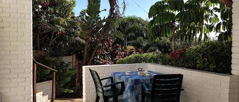 An outdoor patio with a table and chairs, surrounded by lush greenery and a clear blue sky.