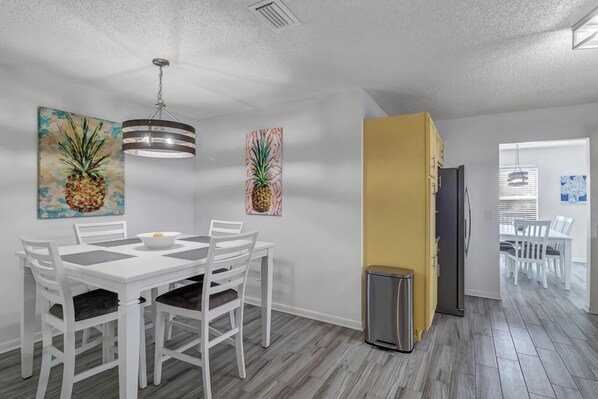 Uma sala de jantar com uma mesa e cadeiras brancas, um lustre pendente e duas pinturas de abacaxi na parede.