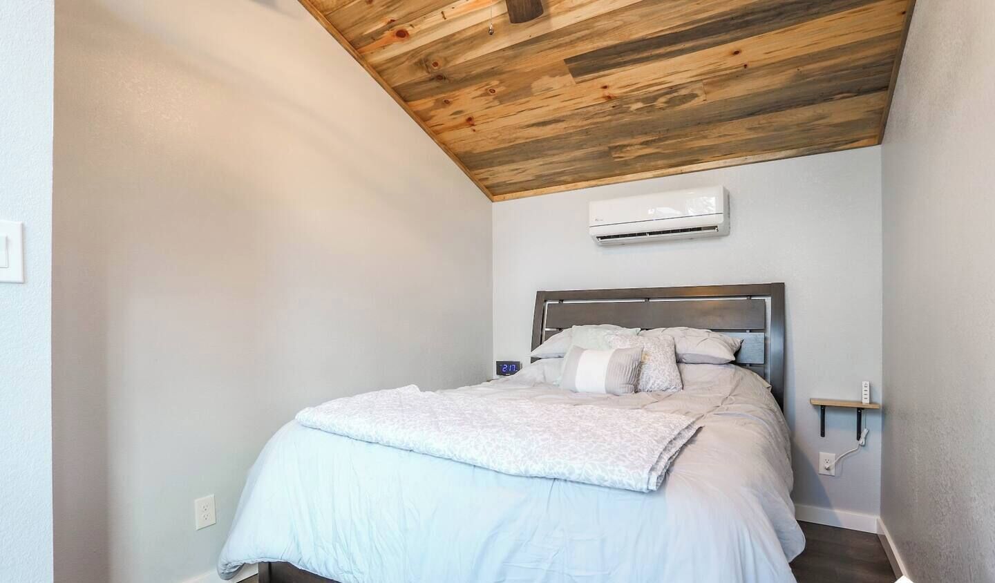 A bedroom with a wooden ceiling, a bed with white bedding, and an air conditioning unit on the wall.