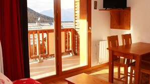 A room with a bed, a wooden table, and a dining table with chairs. There is a sliding glass door leading to an outdoor area with a view of mountains.