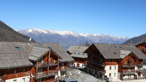A cluster of wooden buildings with balconies, set against a backdrop of snow-capped mountains.