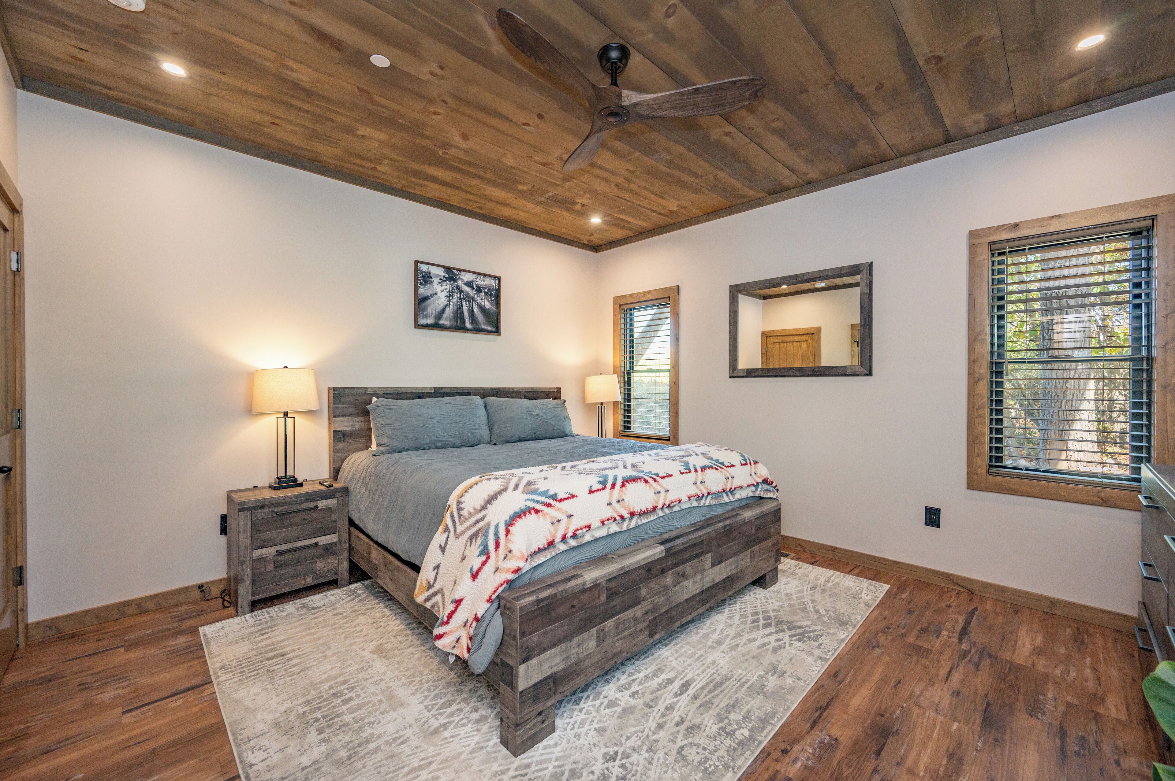 A bedroom with a wooden ceiling, a bed with a patterned blanket, two bedside lamps, and a window with blinds.