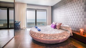 Ein modernes Schlafzimmer mit einem bogenförmigen Bett, Meerblick durch große Fenster und einem Balkon mit einem Stuhl.