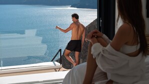 Un hombre con pantalones cortos negros parado junto a una ventana con vista a un cuerpo de agua y montañas al fondo.
