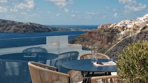 Una mesa con sillas de mimbre y un tablero de vidrio con vistas a un paisaje costero con edificios blancos y acantilados.