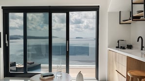 Una habitación moderna con un amplio ventanal corredera que da a un balcón con vistas al mar. Hay una pequeña mesa redonda con un libro y un vaso de agua, y una silla blanca.