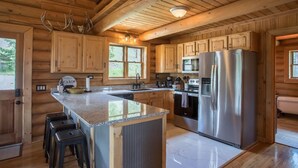 A cabin-style kitchen with wooden cabinets, a stainless steel refrigerator, a granite countertop, and a large island with bar stools. - Remote Mountain Cabin with Incredible Views (Superior)