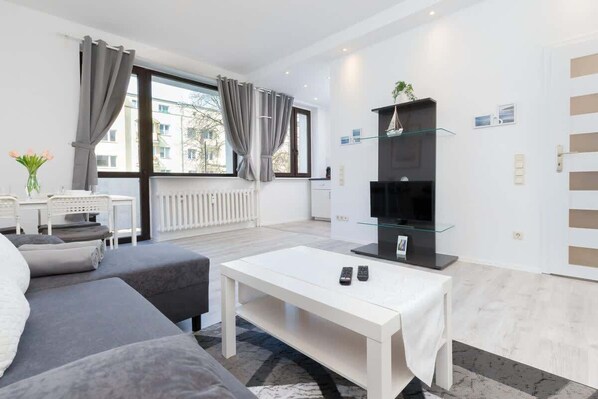 A modern living room with a sofa, a white coffee table, a TV on a glass shelf, and a view of the city through large windows.