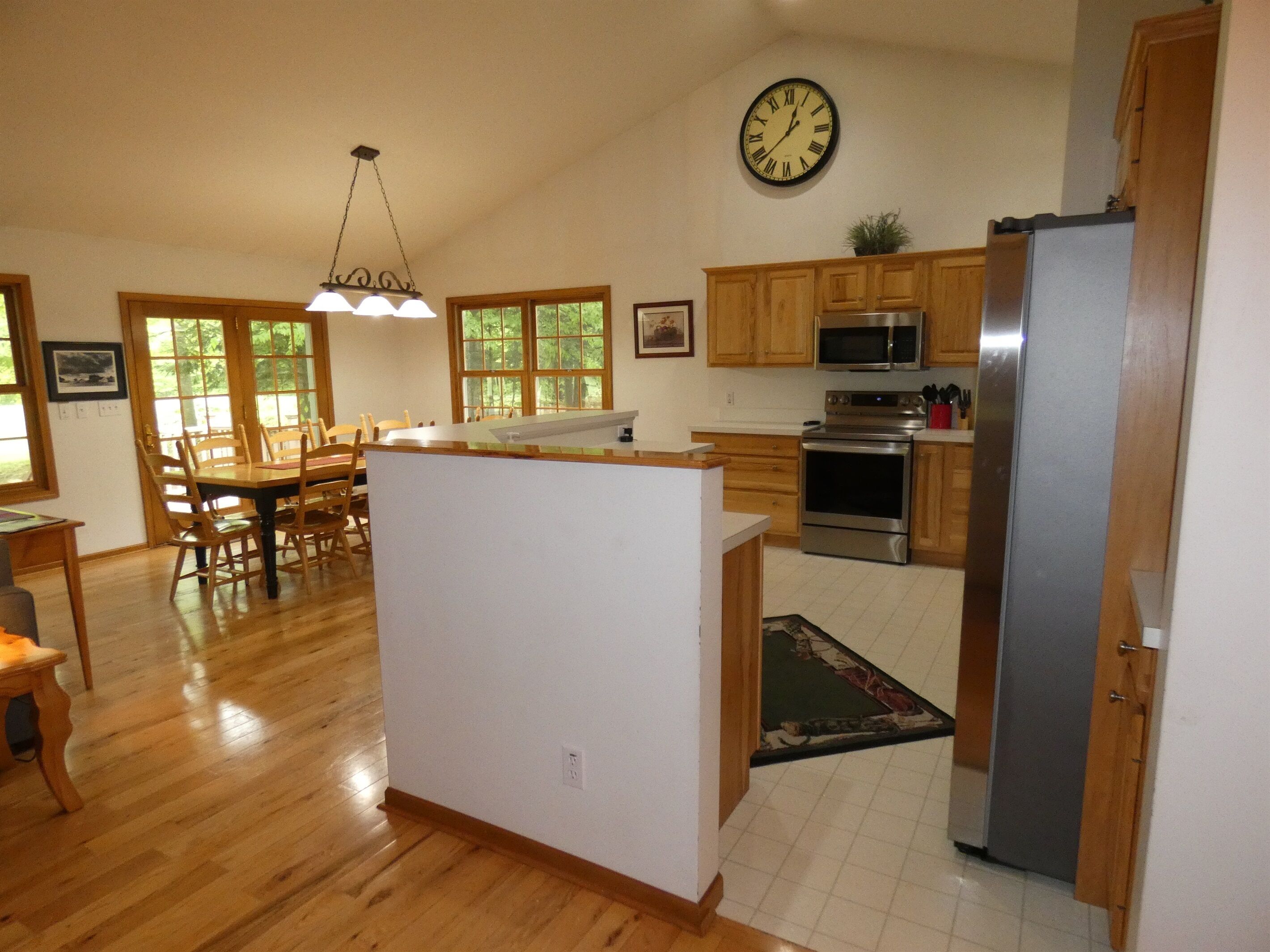 A spacious kitchen with wooden cabinets, a stainless steel refrigerator, a microwave, and an oven. There is a dining area with a table and chairs, and a large window providing natural light.