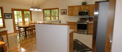 A spacious kitchen with wooden cabinets, a stainless steel refrigerator, a microwave, and an oven. There is a dining area with a table and chairs, and a large window providing natural light.