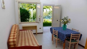 A room with a checkered sofa, a dining table with chairs, a window with a view of greenery, and a door leading outside. - House (Tropea)