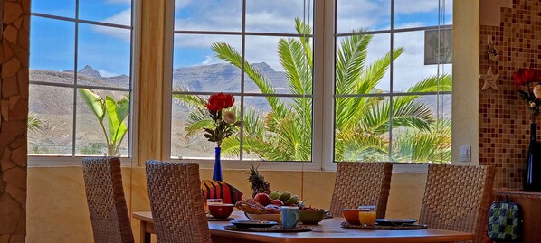 A dining area with wicker chairs, a table with fruits and a vase of flowers, and a view of mountains through large windows.
