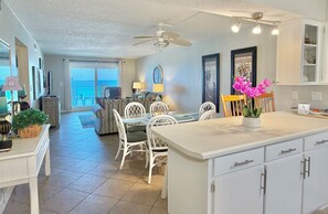 A spacious living area with a dining table, white chairs, a ceiling fan, and a view of the ocean.