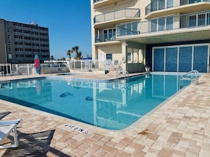 Piscina com placa de 'Não Mergulhar', espreguiçadeiras e um edifício com varandas ao fundo.