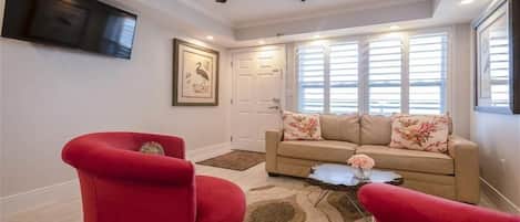 Una sala de estar con un sofá color beige, sillones rojos, un ventilador de techo, un televisor de pantalla plana y un ventanal con persianas.