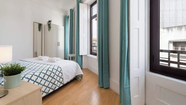 A bedroom with a bed, a bedside table with a plant, a window with curtains, and a view of the outside.