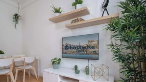 A modern living room with a flat-screen TV mounted on the wall displaying 'hostwise'. There are wooden chairs and a small dining table, along with potted plants on shelves and a floor lamp.