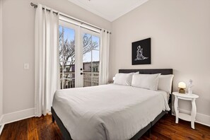 A bedroom with a large bed, white curtains, a small bedside table with a lamp, and a framed picture on the wall. - Perfect Modern Getaway with Parking Steps to Forsyth by Lucky Savannah (Savannah)