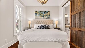 A bedroom with a large bed, a headboard, two bedside tables with lamps, a window with blinds, and a wooden door. - Perfect Modern Getaway with Parking Steps to Forsyth by Lucky Savannah (Savannah)
